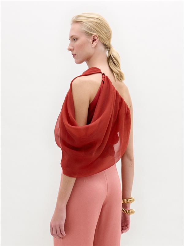 A woman wears the Adil Blouse in red with a one-shoulder neckline, paired with pink high-waisted pants, a wide belt with a large gold buckle, and gold bracelets, standing against a plain white background.
