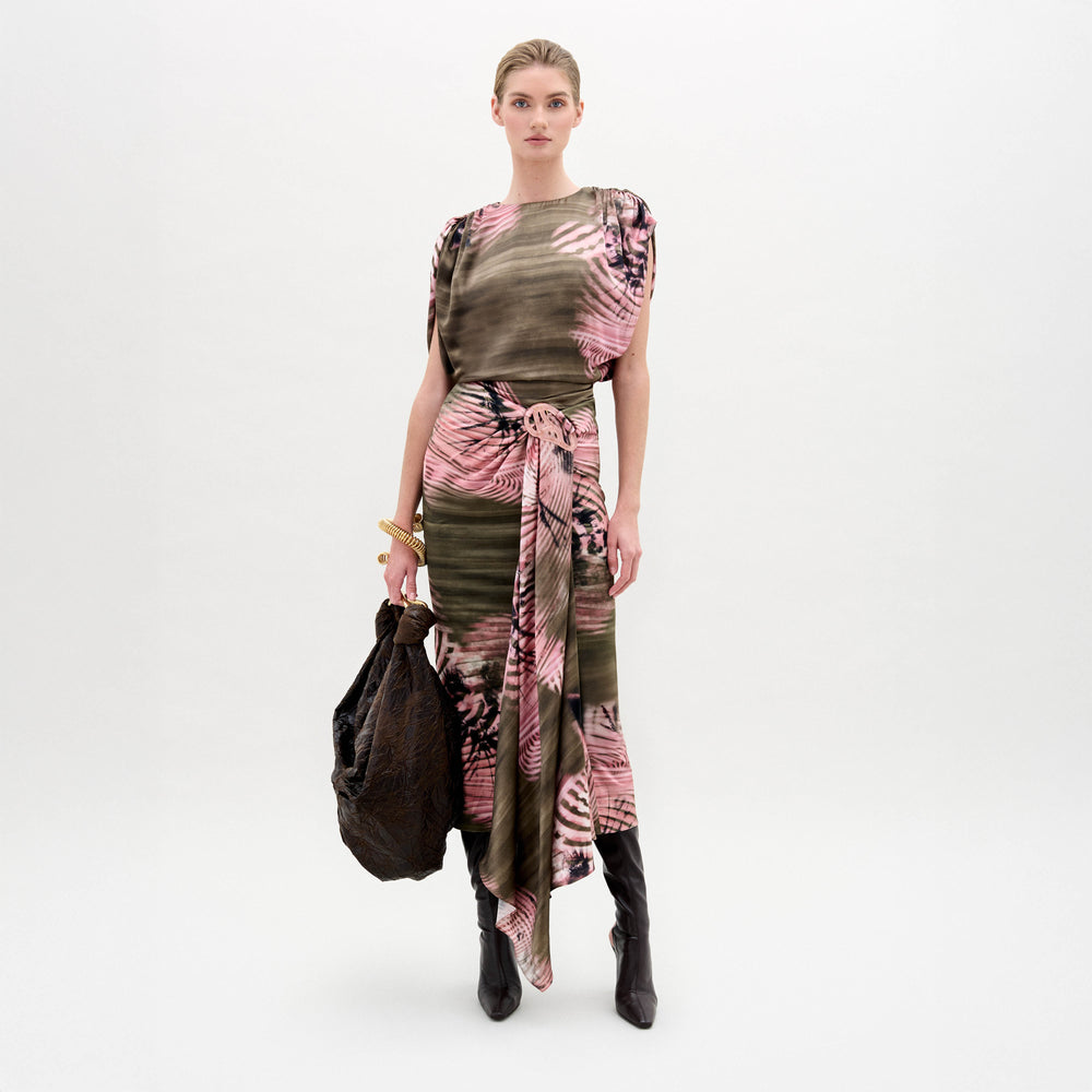 A woman stands against a plain background wearing a pink and brown patterned top, the Adrika Skirt in Italian fabric, black boots, and holding a large brown textured bag.