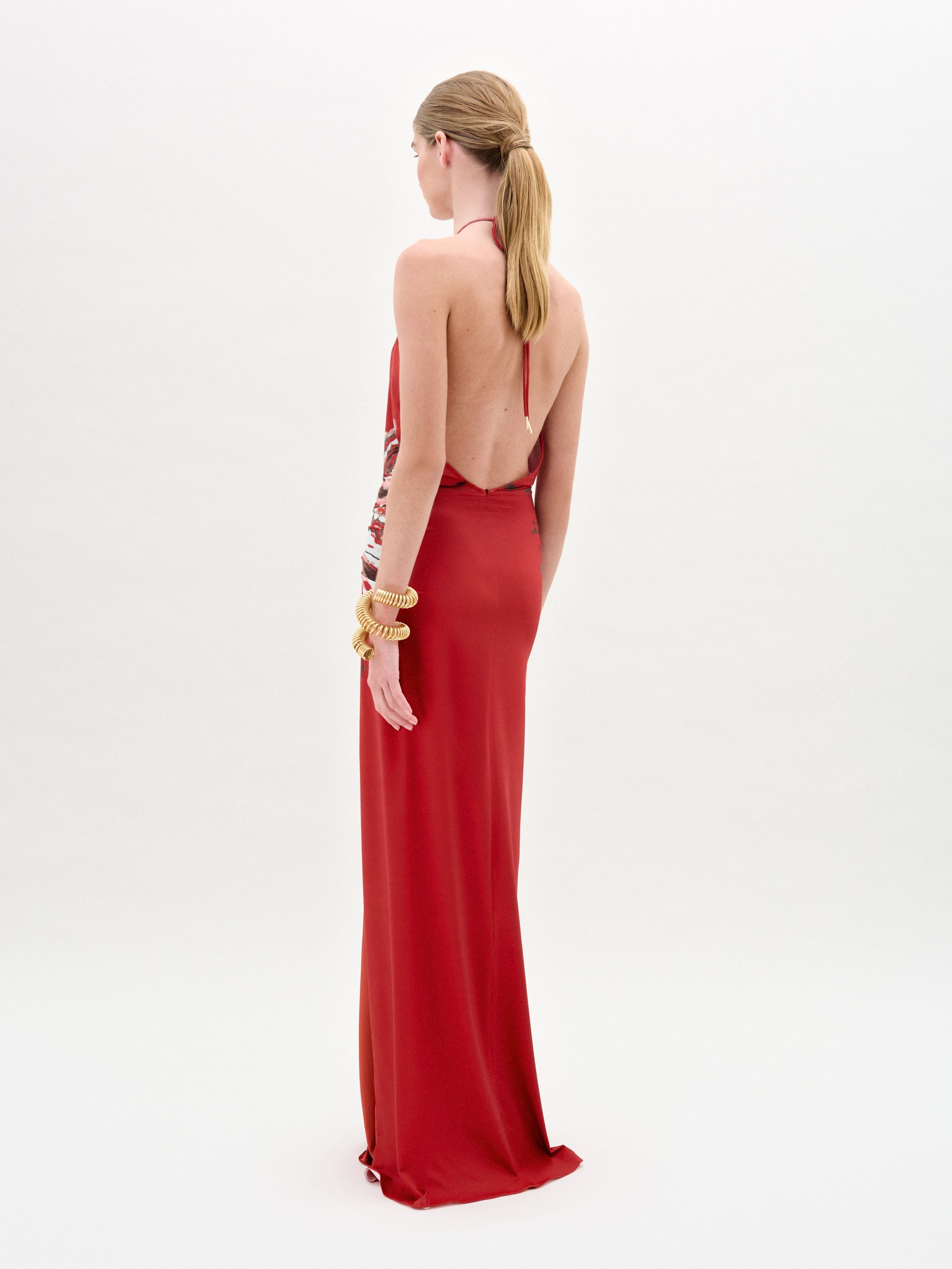 A woman wearing the Aiko Dress—a sleeveless red maxi with a bold white and black burst pattern at the waist and gold embellishments—stands against a plain white background, accessorized with multiple gold bracelets.