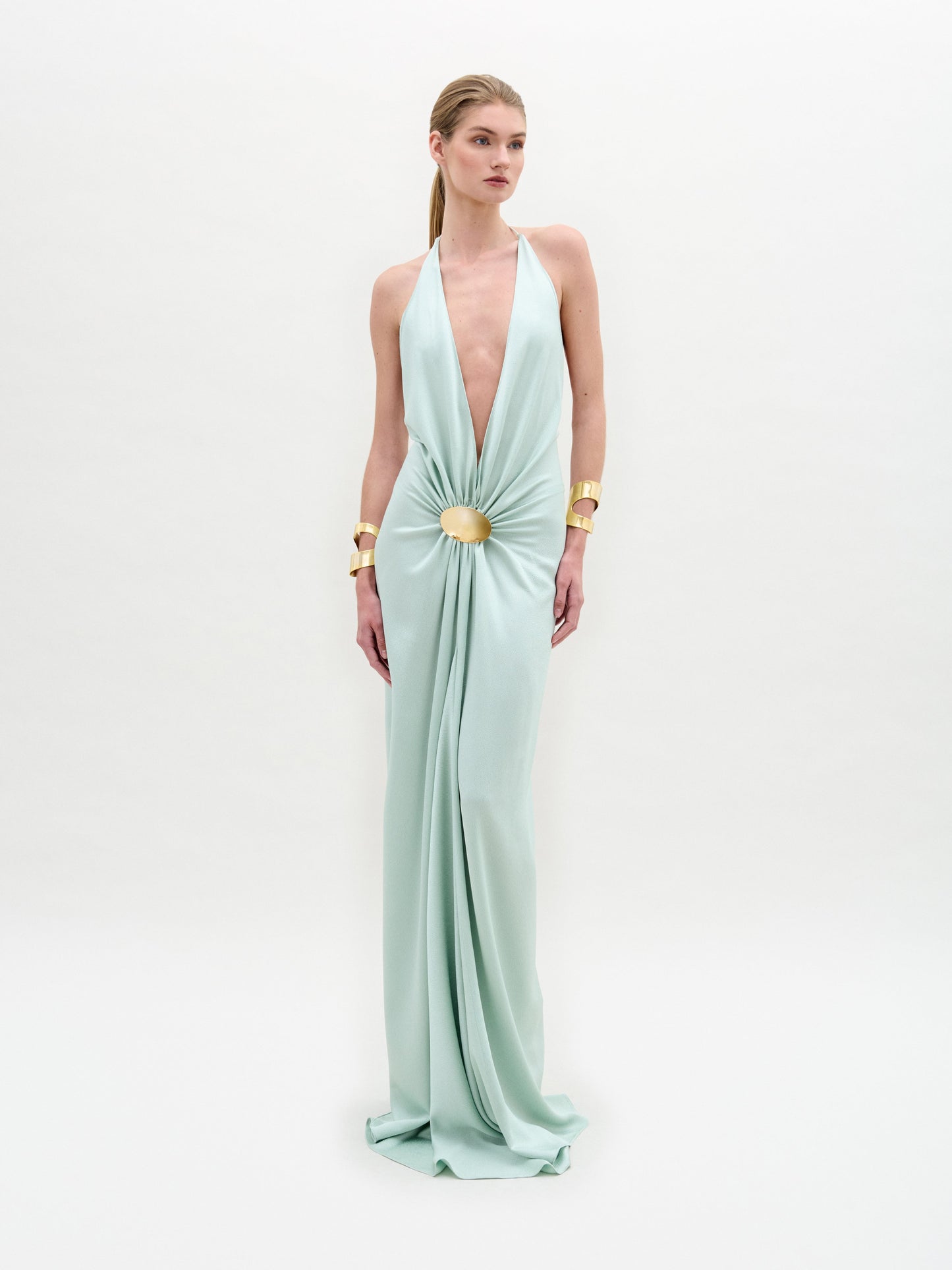 A person wearing the Aiko Dress, a light green halter gown featuring a central gold accent and gold wrist cuffs, stands against a white background.