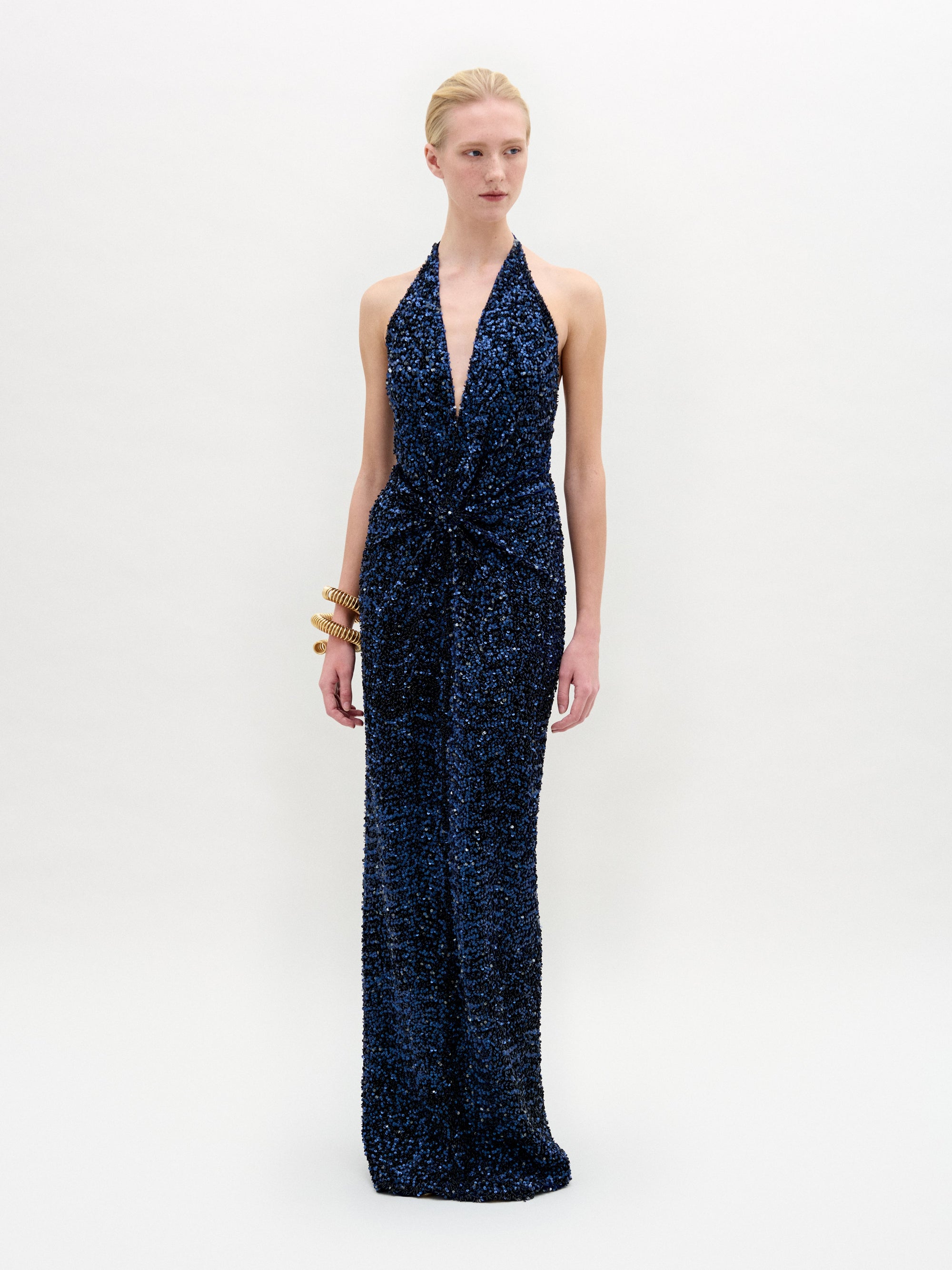 A person dressed in a sleeveless Aiko Dress—a deep blue Atelier creation adorned with sequins and featuring an elegant halter neckline—is accessorized with gold bangles while standing against a plain background.