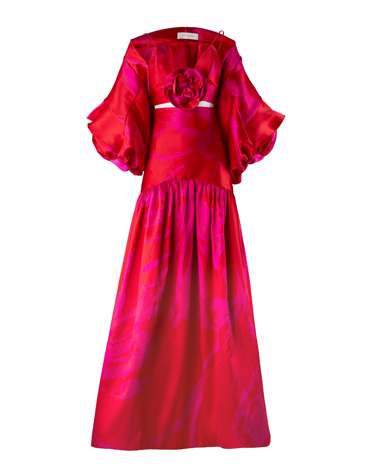 A person with fair skin and dark hair is wearing the Aleen Shrug Rouge Fuchsia Flower, a red, floral-themed outfit featuring large, petal-like sleeves and a central rosette. Set against a plain white background, this stunning piece is available for pre-order now and will ship by February 2025.