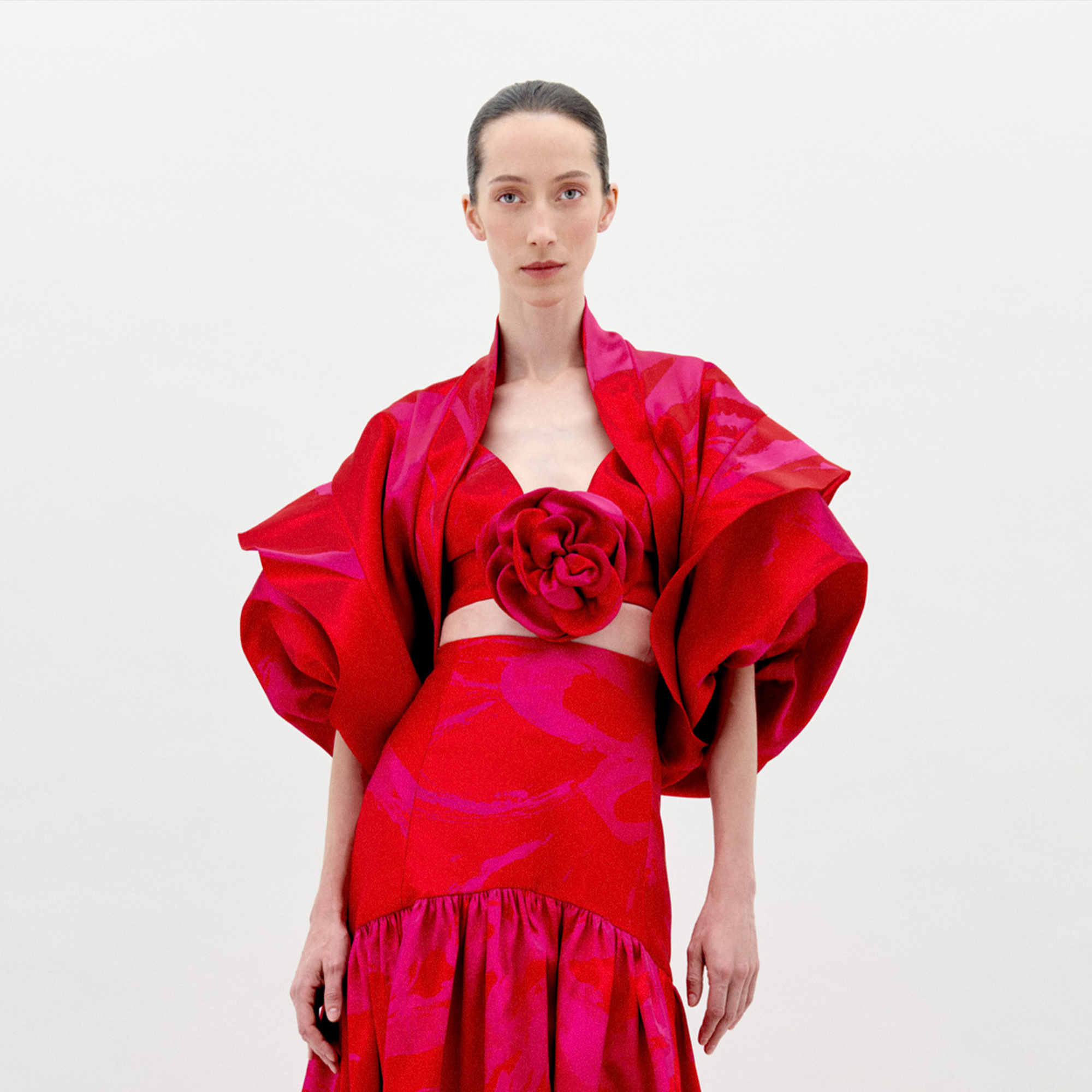 A person with fair skin and dark hair is wearing the Aleen Shrug Rouge Fuchsia Flower, a red, floral-themed outfit featuring large, petal-like sleeves and a central rosette. Set against a plain white background, this stunning piece is available for pre-order now and will ship by February 2025.