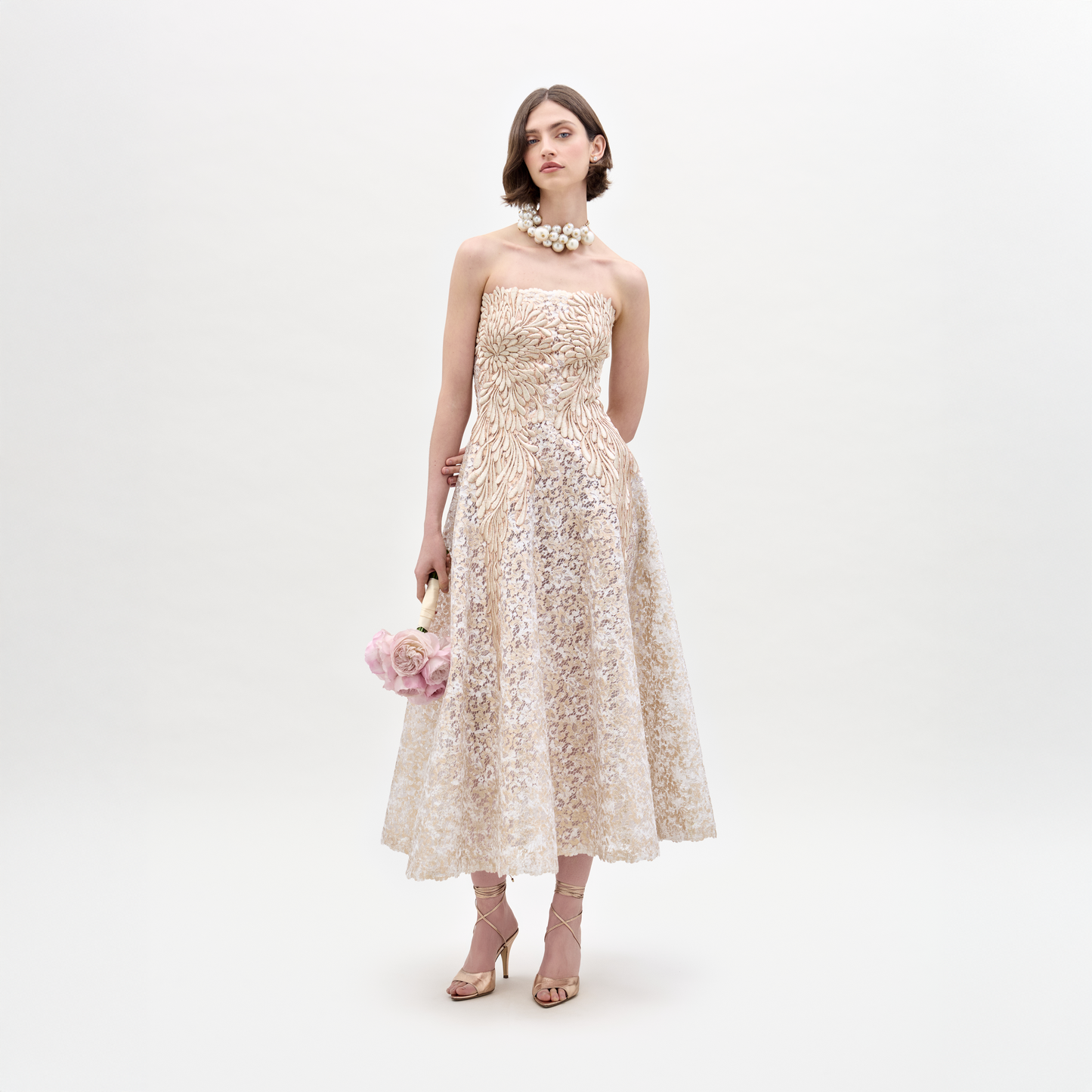 A woman in the strapless Alejandra Dress holds pink flowers. She wears pearl jewelry and beige heels, standing against a plain white background.