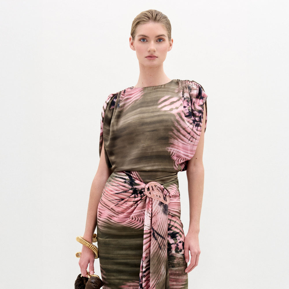 A woman stands against a plain background wearing the Alisa Blouse, featuring a pink and olive green pattern crafted from Italian fabric. She holds a brown bag and wears gold bracelets.