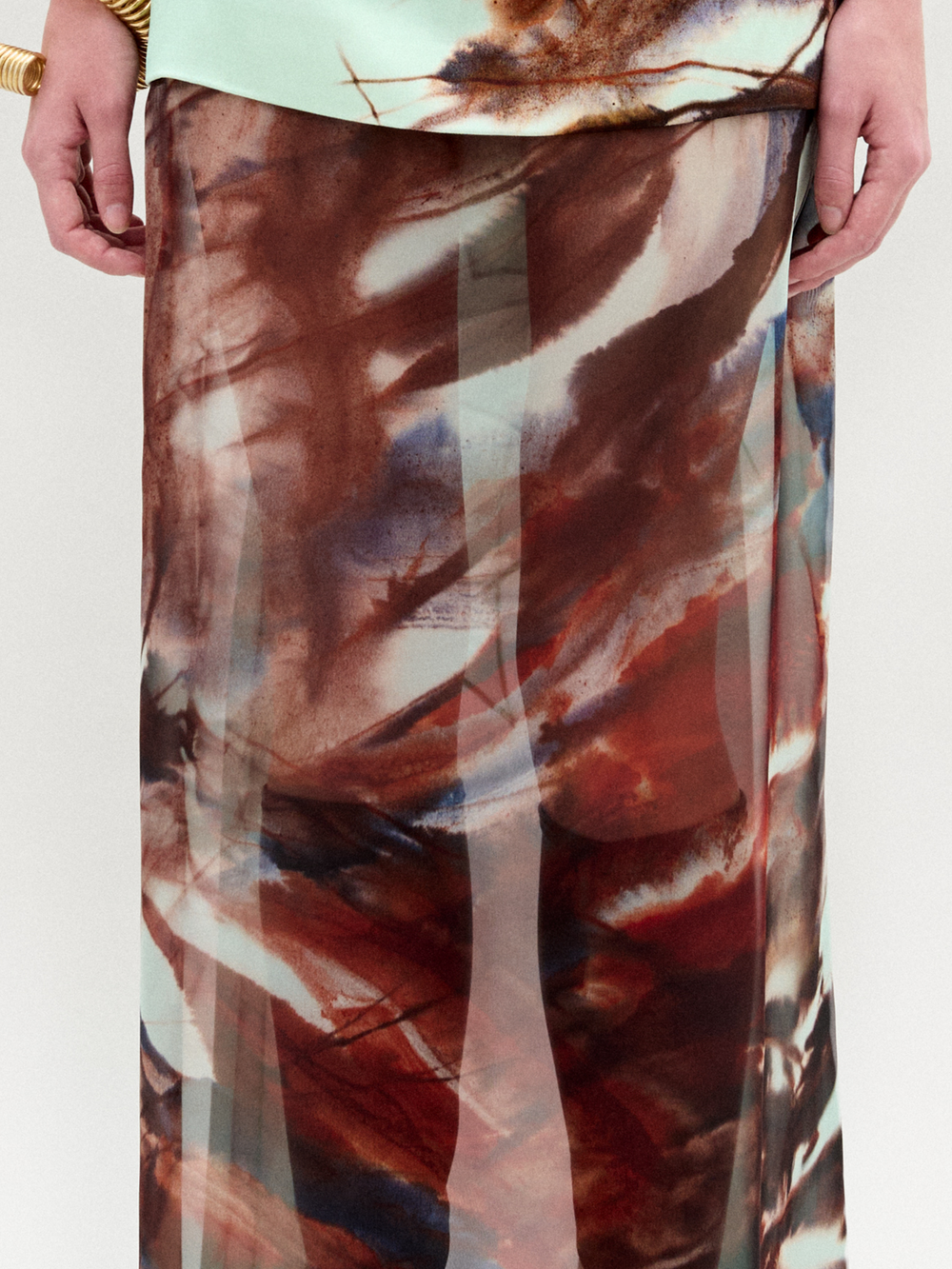 A woman poses against a plain backdrop in the Amalia Skirt—a flowing maxi with an abstract brown and blue pattern—paired with a sleeveless halter top, wide black belt, and bracelets. The look features luxurious Italian fabric.