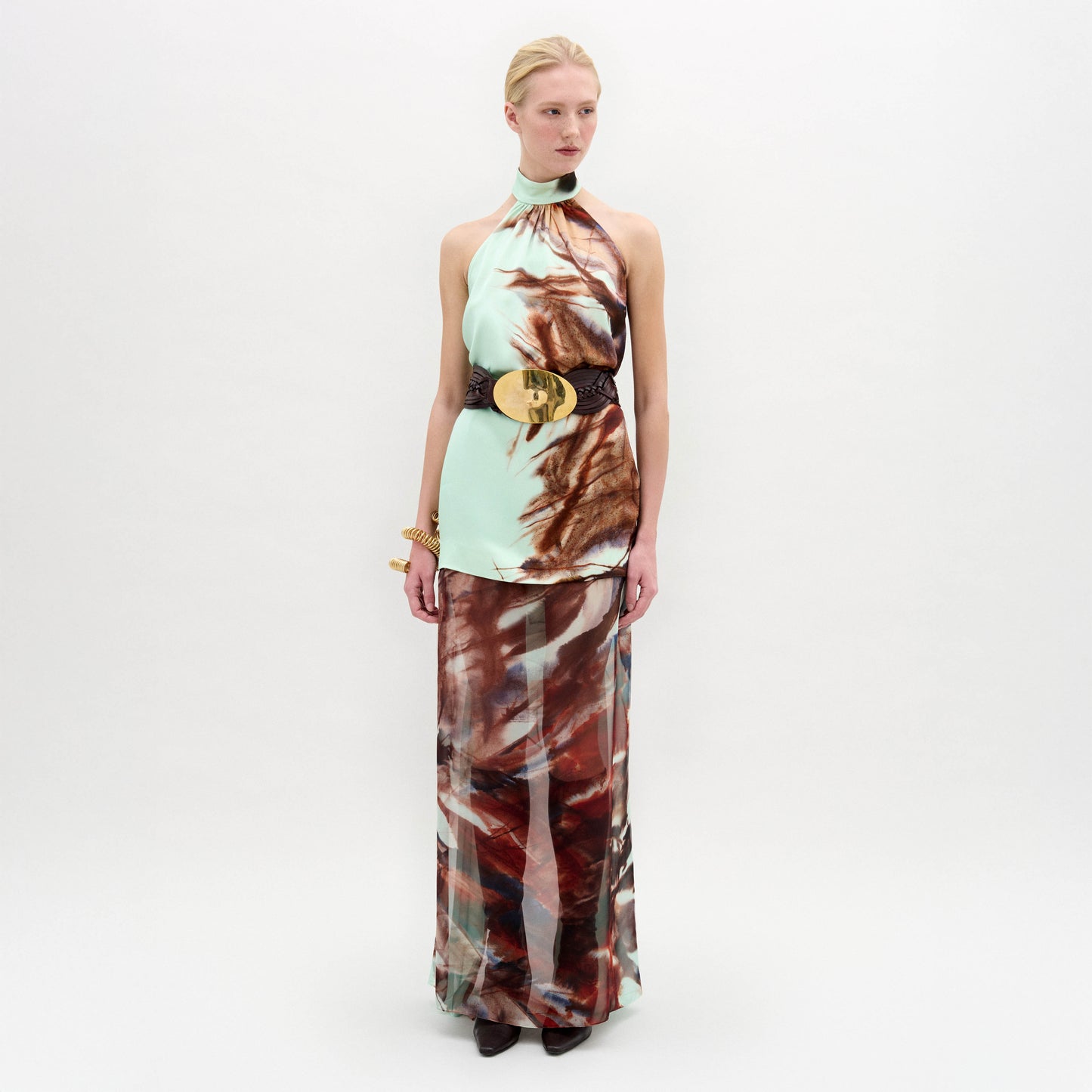 A woman poses against a plain backdrop in the Amalia Skirt—a flowing maxi with an abstract brown and blue pattern—paired with a sleeveless halter top, wide black belt, and bracelets. The look features luxurious Italian fabric.