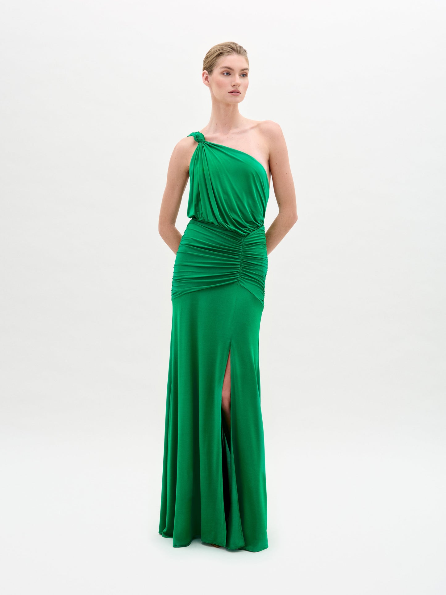 A woman in the Antara Dress, a green one-shoulder gown with a side slit, stands against a plain white background.