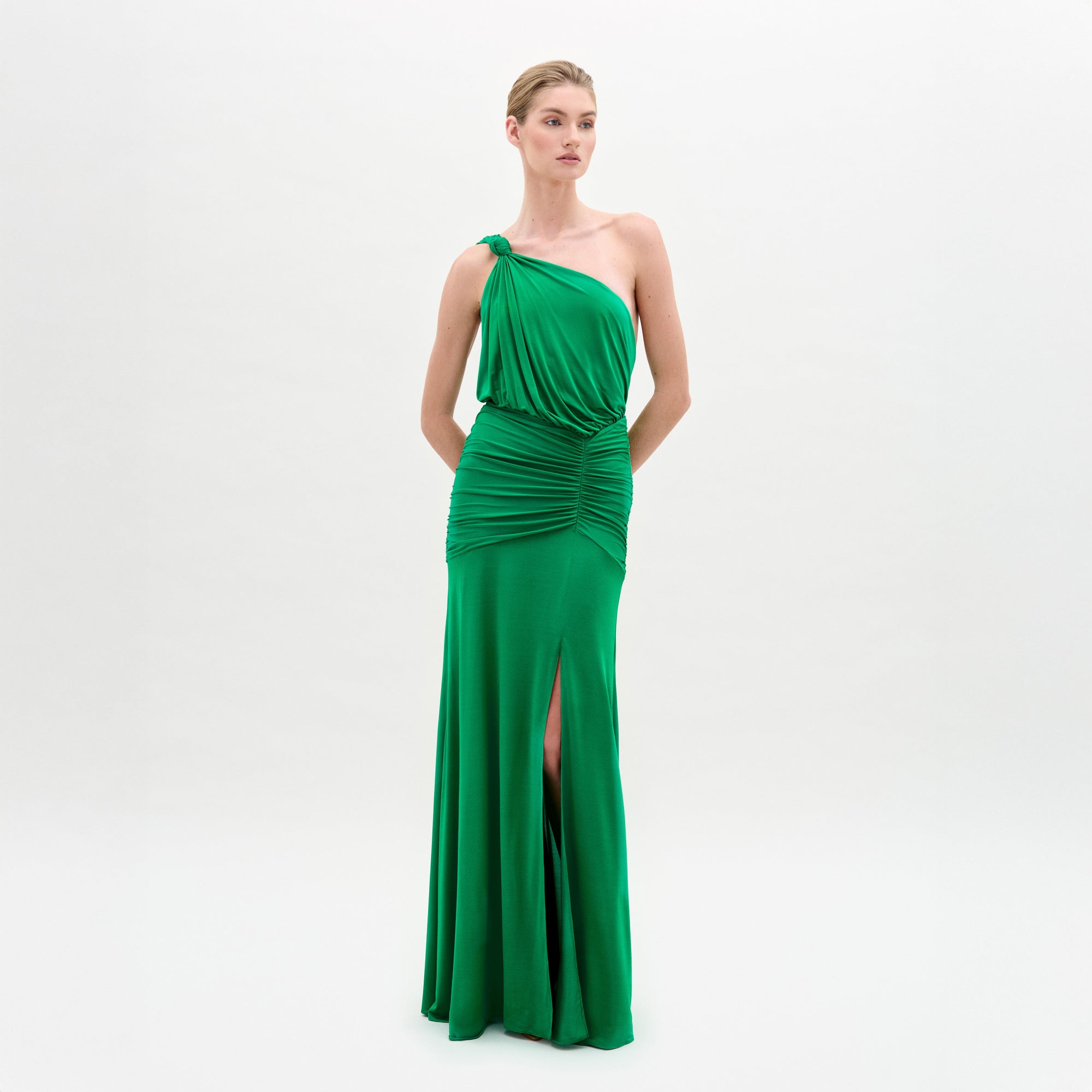 A woman stands against a plain white background wearing the Antara Dress—a bright green, fitted maxi with an asymmetric one-shoulder neckline, gathered waist, and front slit.