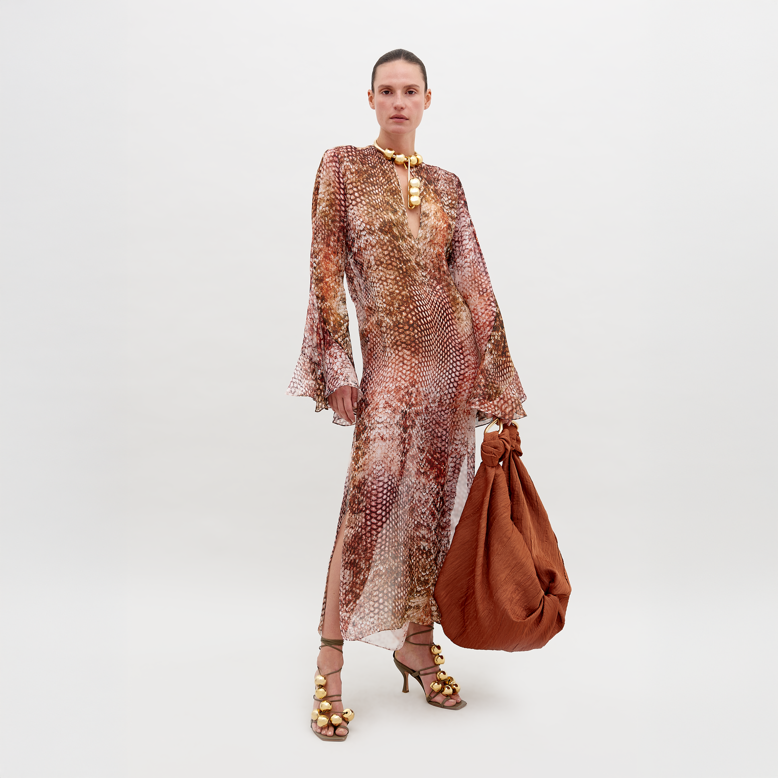 A person stands against a plain background wearing the Aries Dress Peach Animal Print with flared sleeves, embellished accessories, and high-heeled sandals. They hold a large, rust-colored bag. Pre-order now to have it by November 15th, 2024.