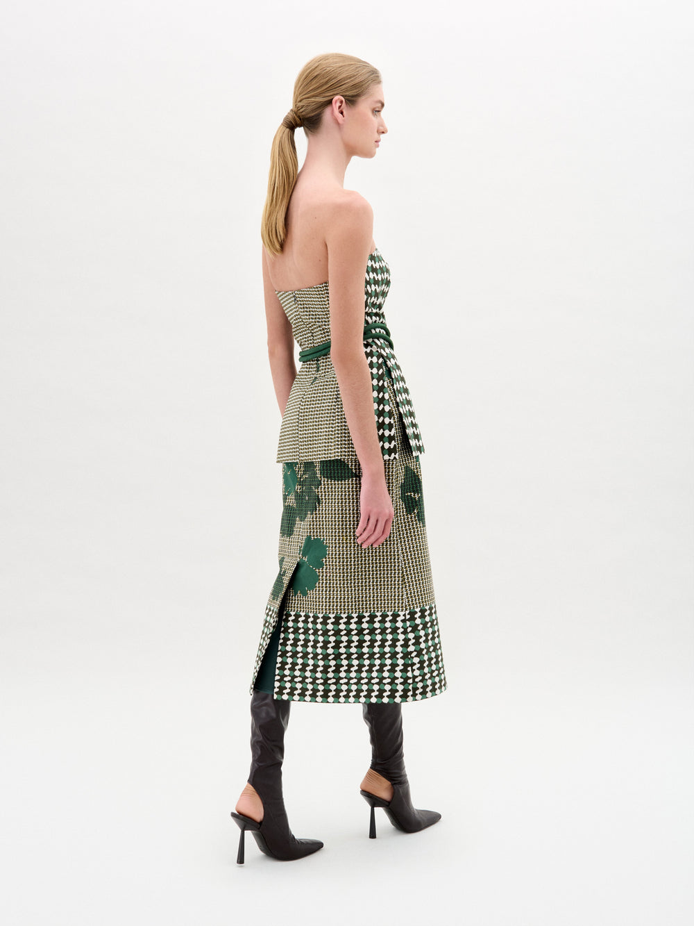 A woman stands against a plain background wearing the Atira Skirt, a green and beige abstract print design with a belted waist, crafted from Italian fabric, paired with knee-high black boots.