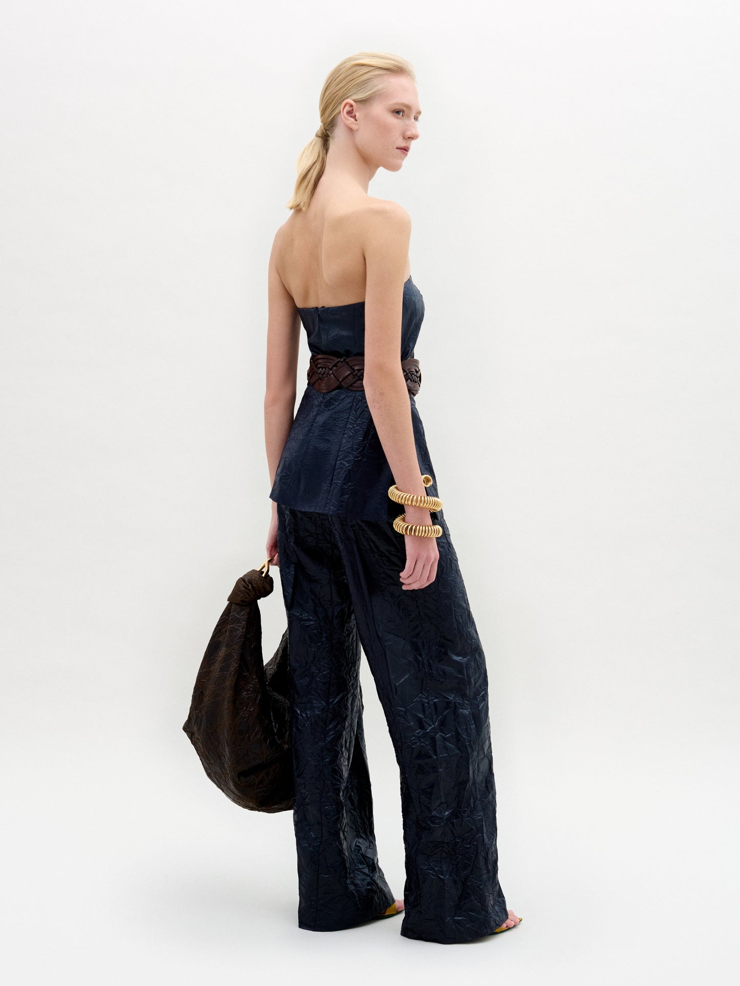 Wearing the Emine Pant, known for its strapless navy design and wide-leg silhouette, the individual holds a large dark jacket over one shoulder. Stylishly accessorized with a wide belt and gold bracelet, they pose against a plain background.