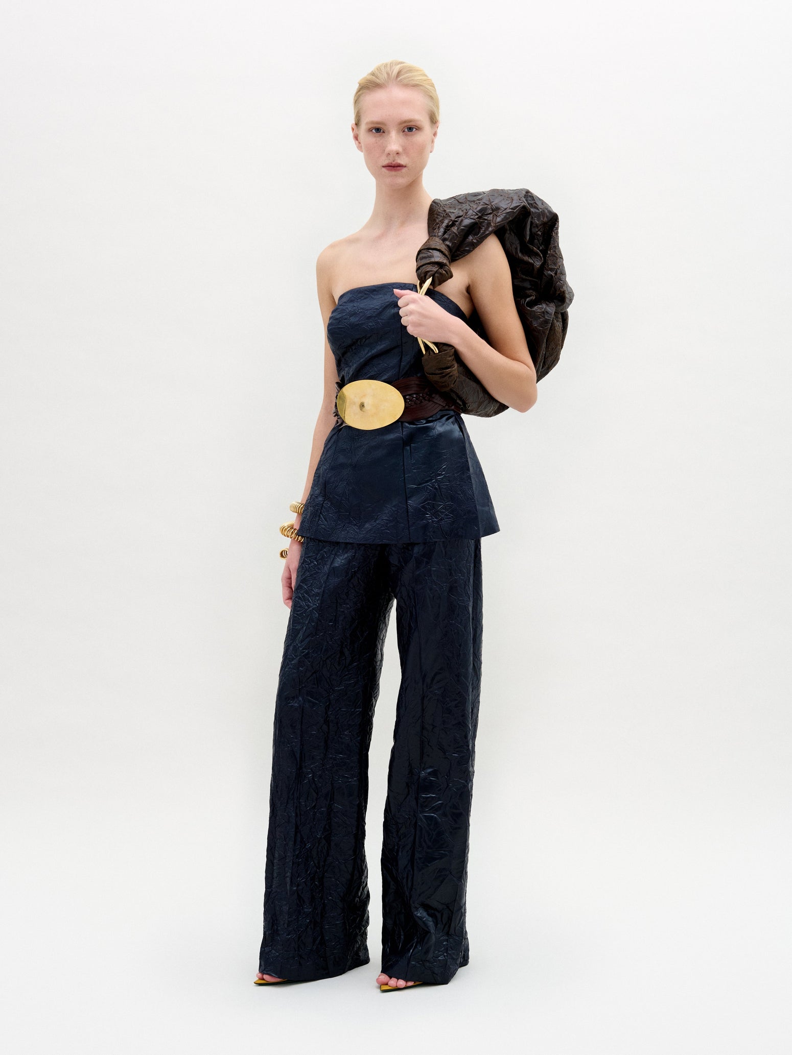 Wearing the Emine Pant, known for its strapless navy design and wide-leg silhouette, the individual holds a large dark jacket over one shoulder. Stylishly accessorized with a wide belt and gold bracelet, they pose against a plain background.