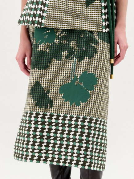 A woman stands against a plain background wearing the Atira Skirt, a green and beige abstract print design with a belted waist, crafted from Italian fabric, paired with knee-high black boots.