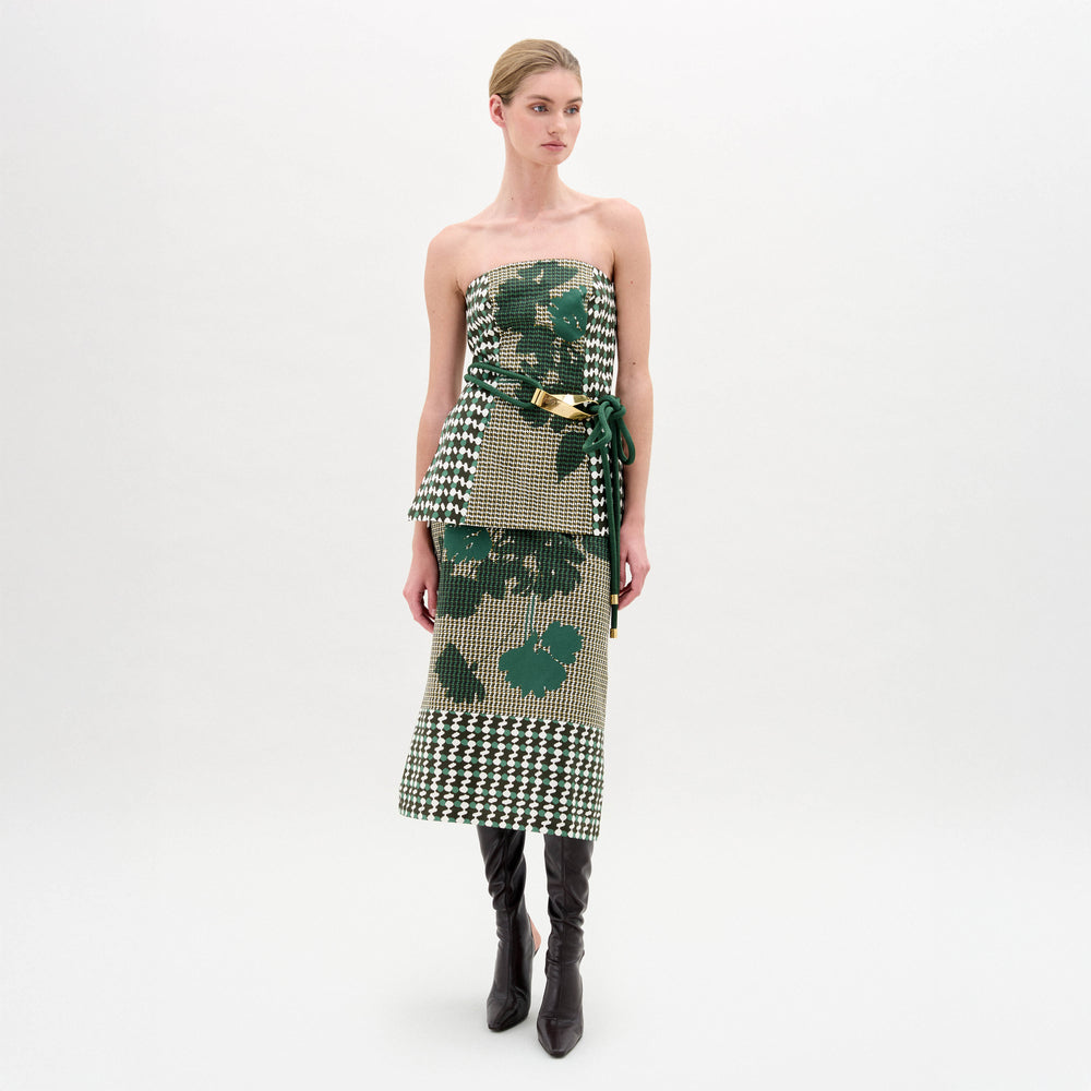 A woman stands against a plain background wearing the Atira Skirt, a green and beige abstract print design with a belted waist, crafted from Italian fabric, paired with knee-high black boots.