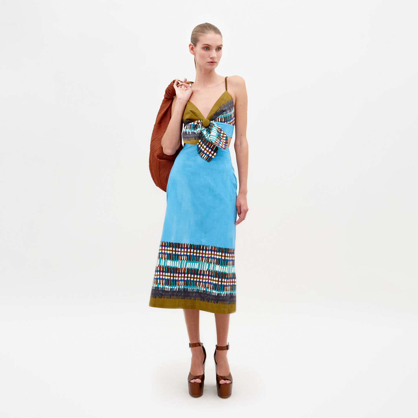 A woman is wearing the Atira Skirt in an aqua olive plaid pattern along with a large bow. She has an orange jacket draped over her shoulder and is standing against a plain white background. This look will be available for pre-order starting February 15th, 2025.