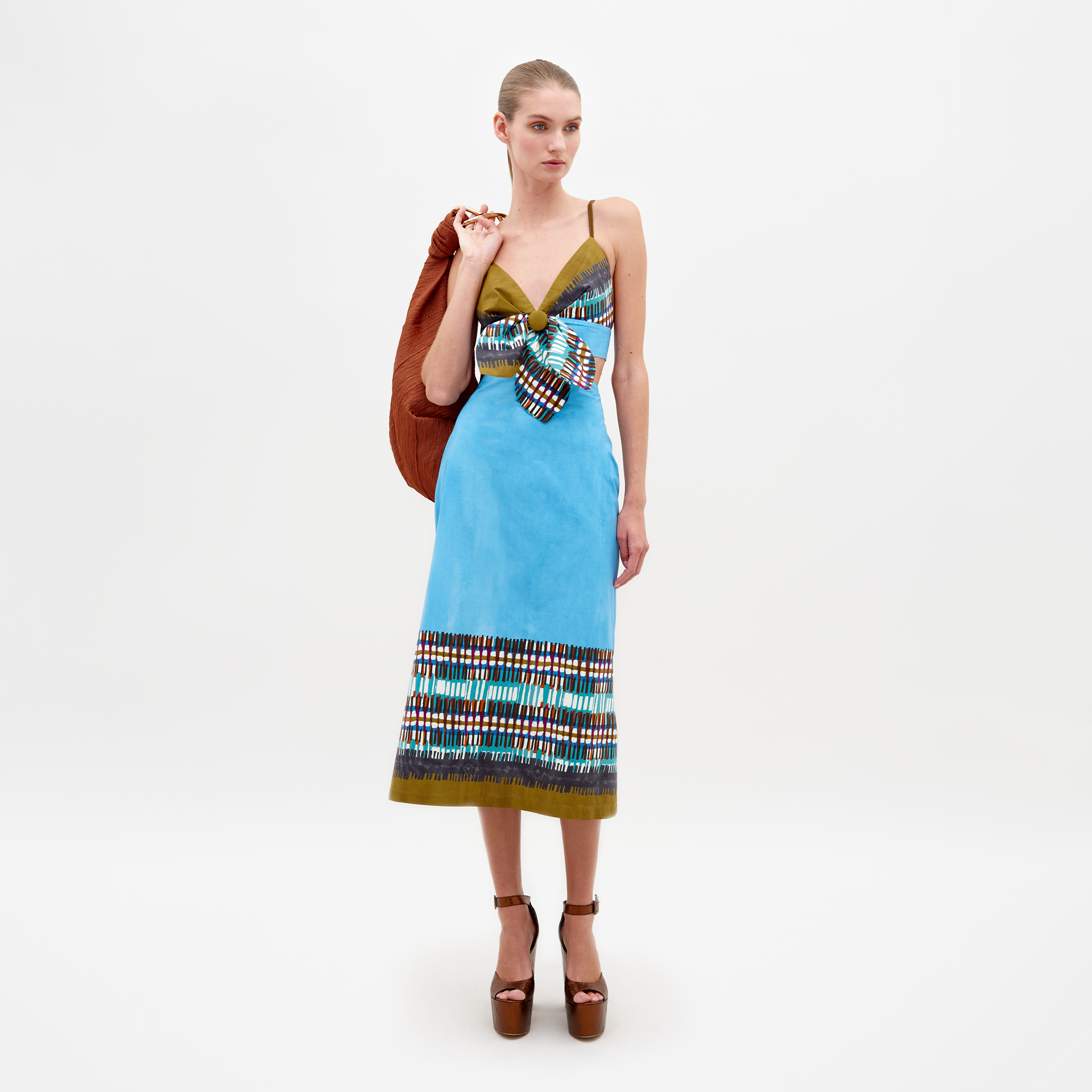 A woman is wearing the Atira Skirt in an aqua olive plaid pattern along with a large bow. She has an orange jacket draped over her shoulder and is standing against a plain white background. This look will be available for pre-order starting February 15th, 2025.