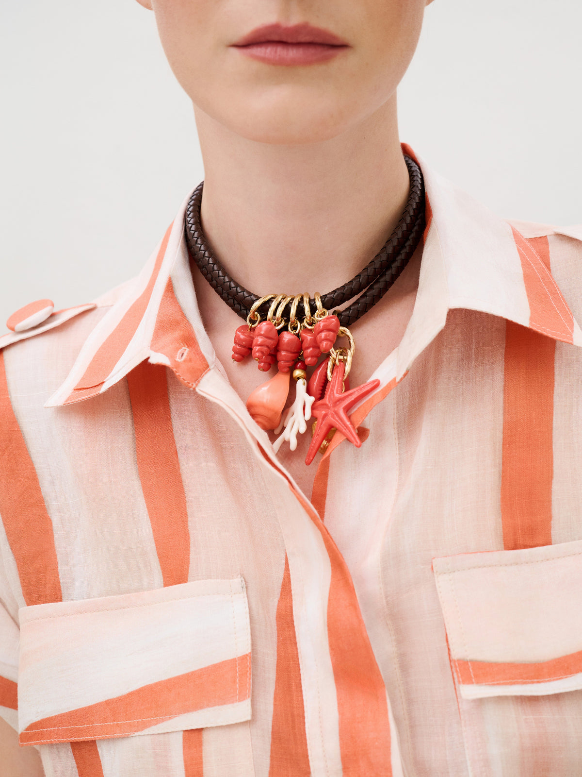 A person wearing an Aaron Romper in Italian fabric and a brown braided leather necklace featuring red and pink charms shaped like coral, shells, and a starfish.