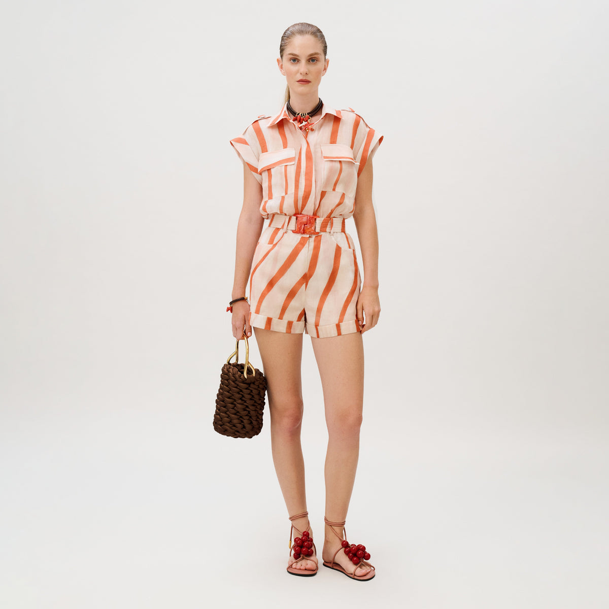 A model wears the orange and white striped linen Aaron Romper with a matching belt, holding a dark brown woven handbag and wearing red-accented lace-up sandals.