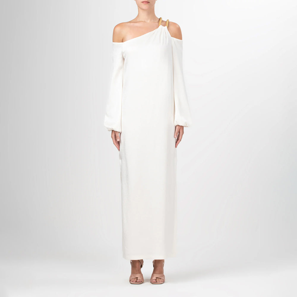 A woman wearing the Ada Dress, a white one-shoulder maxi with an asymmetric strap, stands against a plain light background, paired with peach open-toe heels.