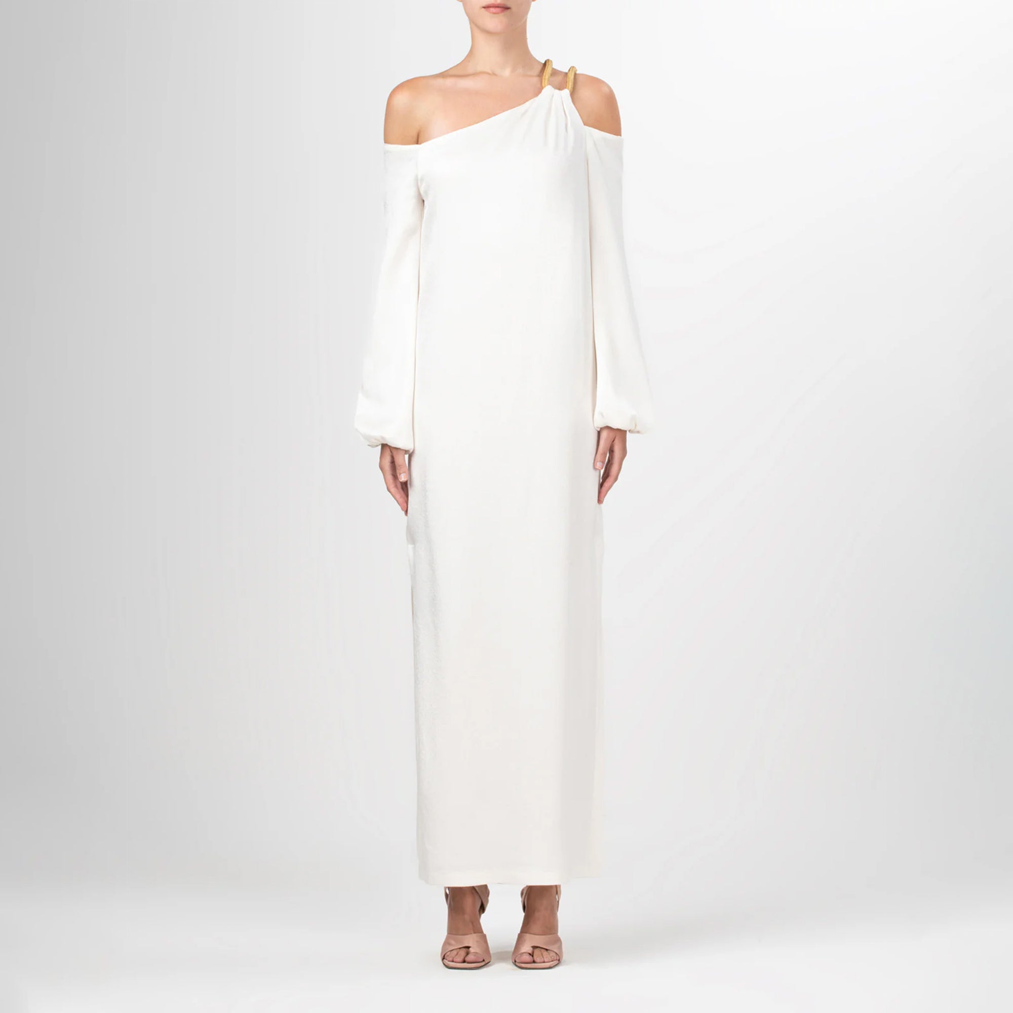 A woman wearing the Ada Dress, a white one-shoulder maxi with an asymmetric strap, stands against a plain light background, paired with peach open-toe heels.