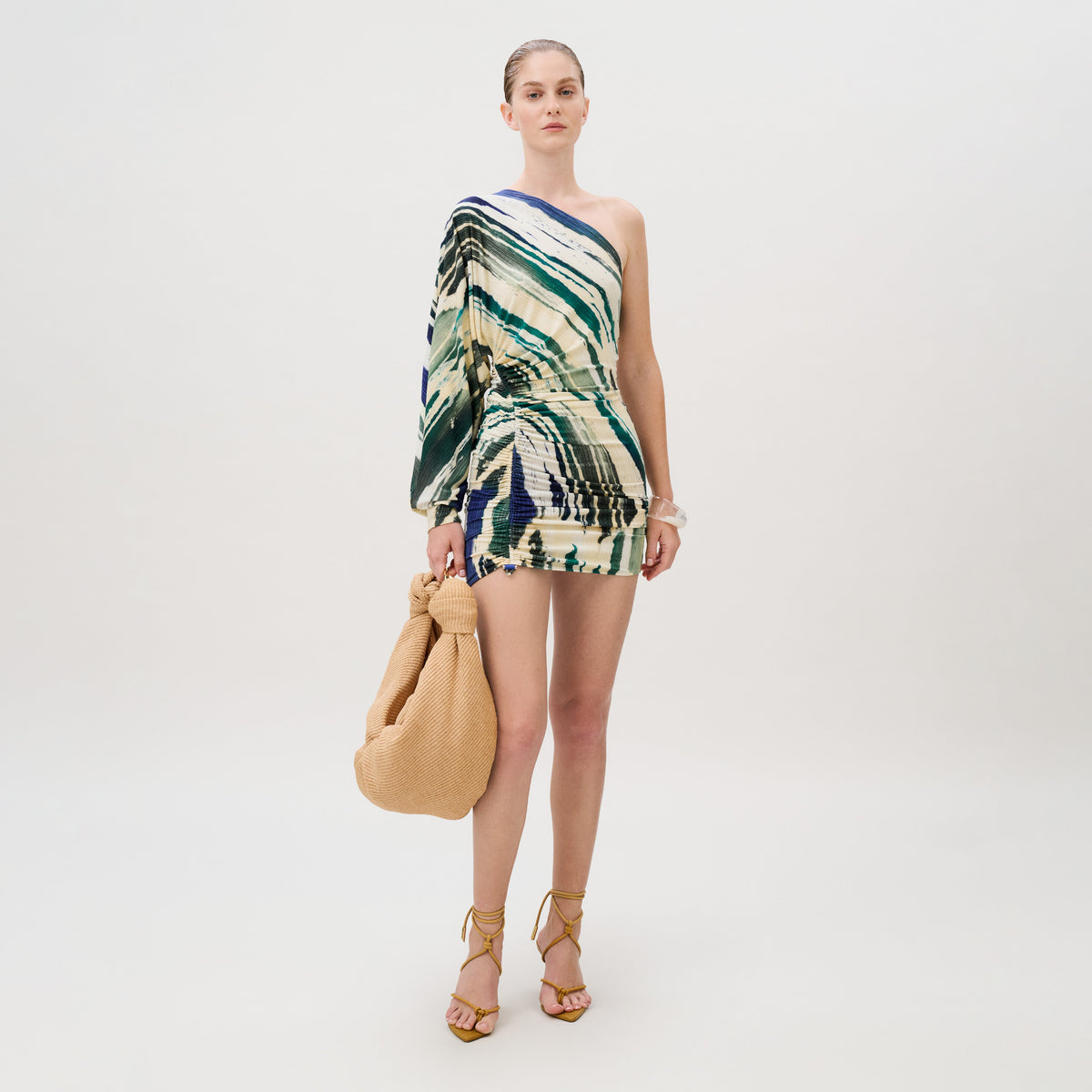 A woman wears the Adelaide Dress, a one-shoulder mini dress in Italian fabric with an abstract green and blue print, carrying a tan woven handbag and wearing strappy high heels.