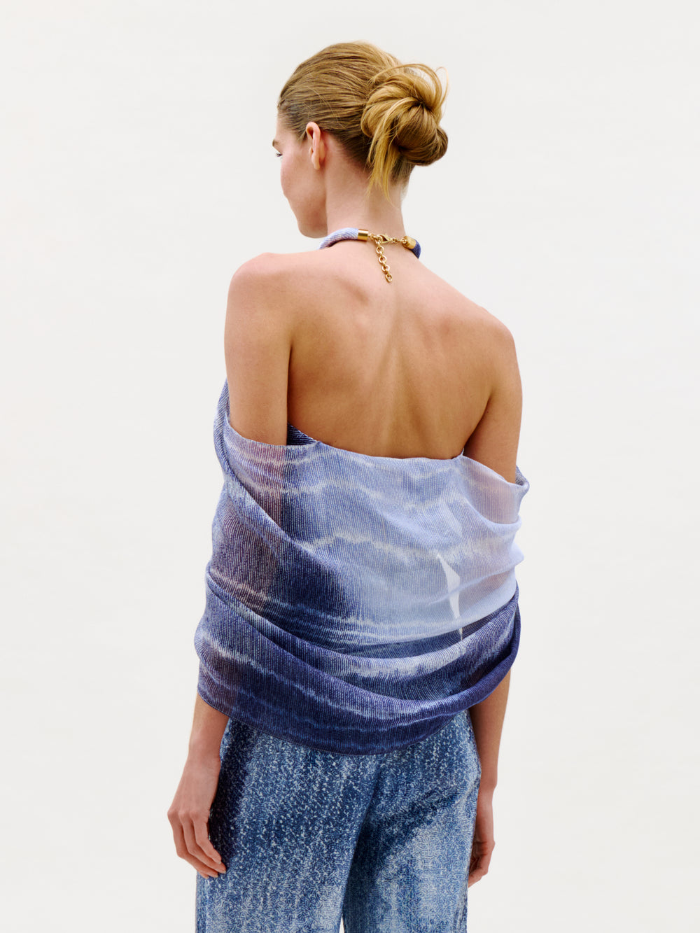 A woman with light brown hair in a bun wears the Adib Blouse—a strapless, blue and white textured wrap top crafted from luxurious Italian fabric—paired with blue patterned pants, standing against a plain, light background and facing away from the camera.
