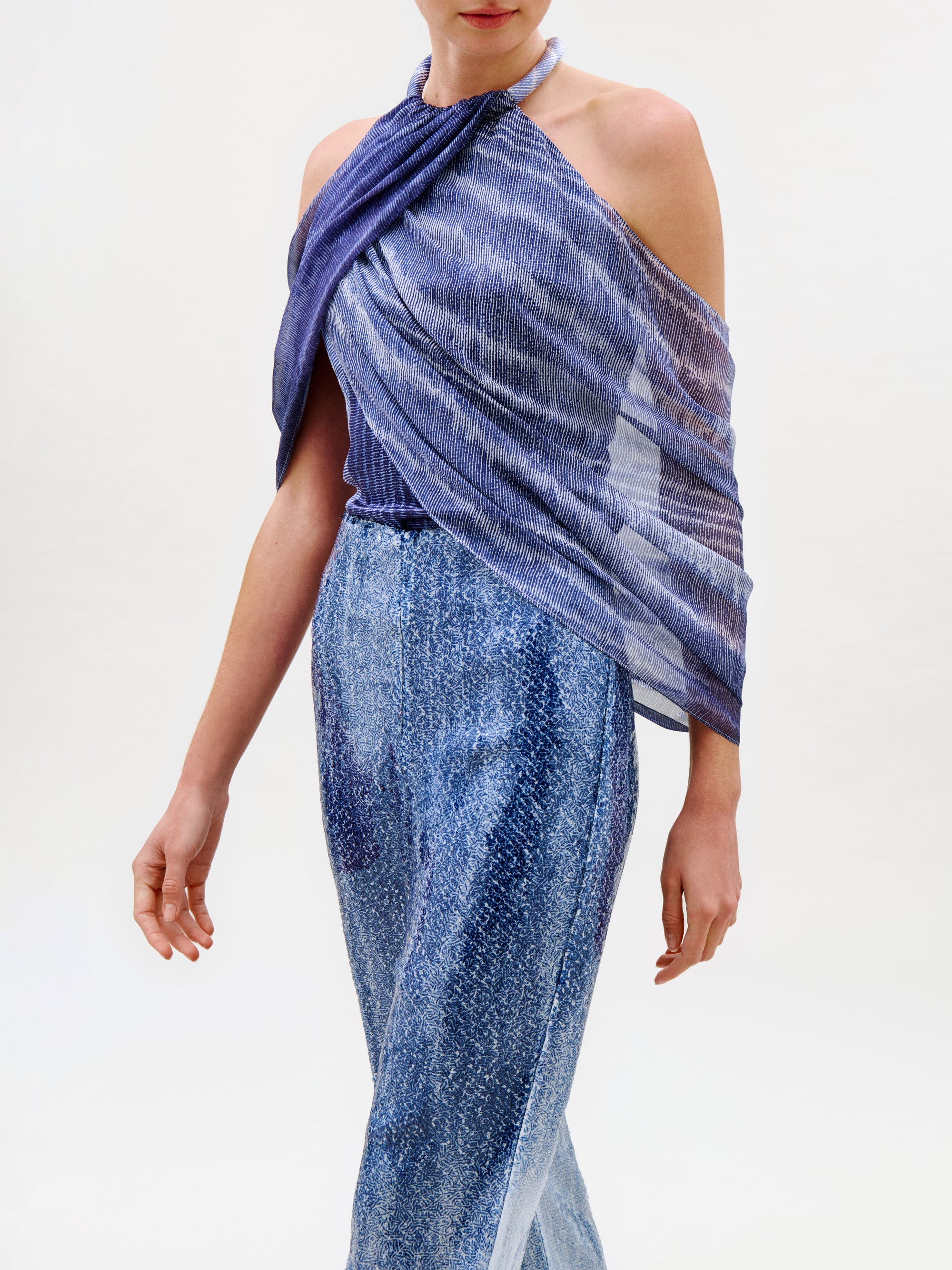A woman wearing the Adib Blouse—a shimmery, crisscross, sleeveless halter top crafted from luxurious Italian fabric in purple—paired with high-waisted metallic blue pants, stands against a plain white background.