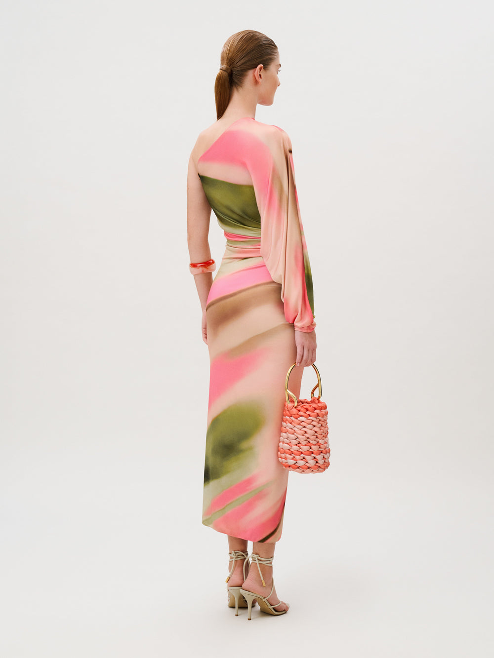 Seen from the back, a person wears the Albany Dress, a pink and olive green abstract print midi dress with a one-shoulder neckline, paired with strappy heels and a braided pink handbag.