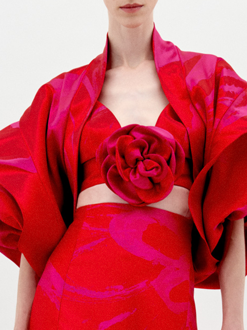 A person with fair skin and dark hair is wearing the Aleen Shrug Rouge Fuchsia Flower, a red, floral-themed outfit featuring large, petal-like sleeves and a central rosette. Set against a plain white background, this stunning piece is available for pre-order now and will ship by February 2025.