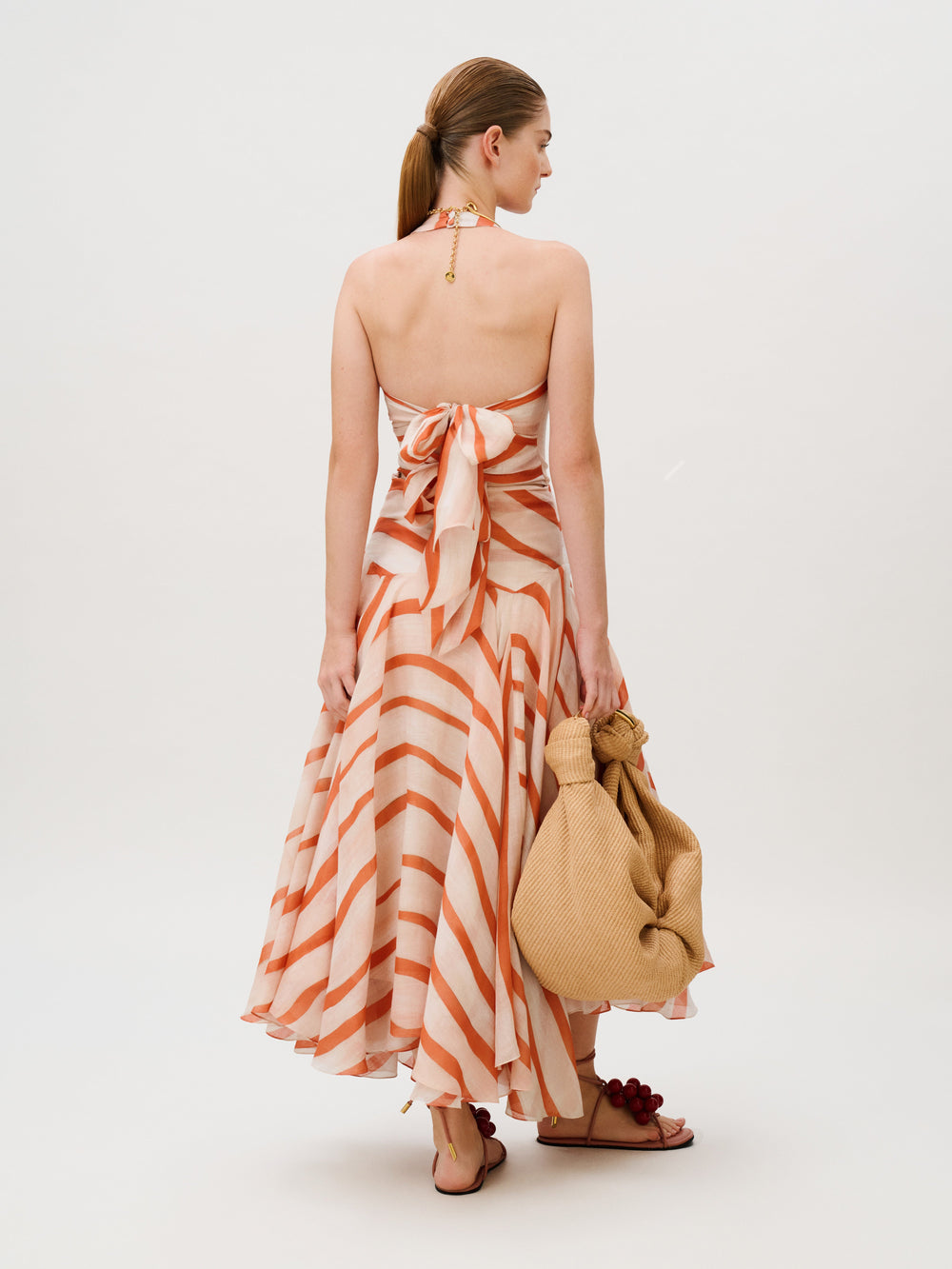 Back view of a person wearing the Amity Dress, a 100% cotton halter-neck midi dress featuring orange and cream diagonal stripes, while holding a beige woven bag.