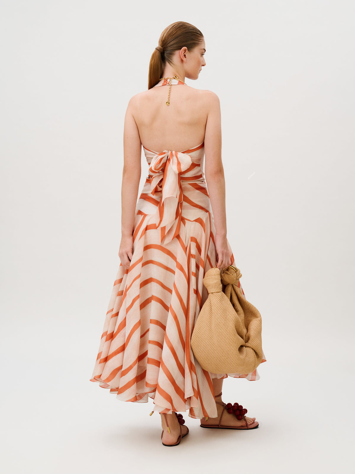 Back view of a person wearing the Amity Dress, a 100% cotton halter-neck midi dress featuring orange and cream diagonal stripes, while holding a beige woven bag.