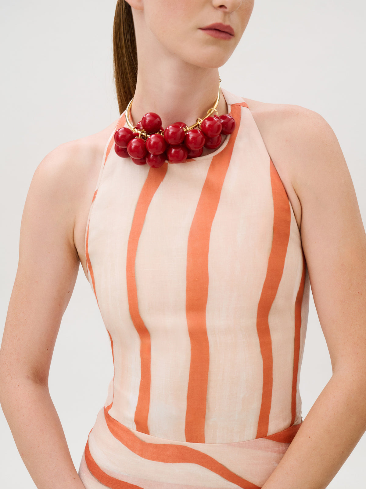 The Amity Dress is a sleeveless, 100% cotton halter dress in orange and cream stripes, worn with a large red cherry-shaped statement necklace.