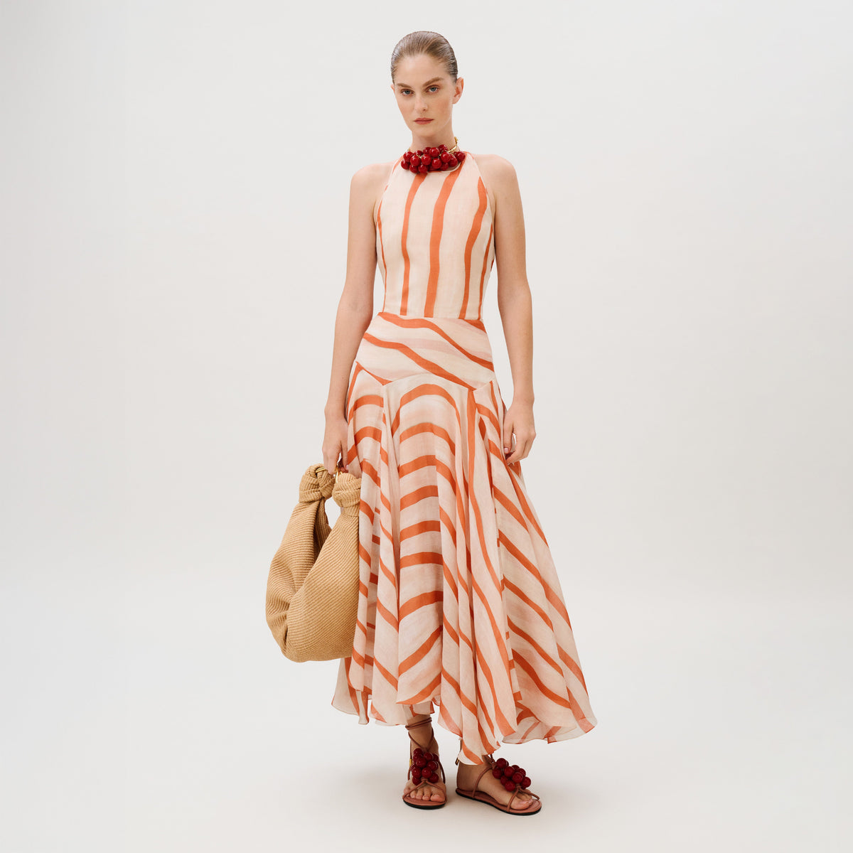 A model wears the Amity Dress, a sleeveless, halter-neck maxi in 100% cotton with orange and white diagonal stripes, paired with a woven tote bag and embellished flat sandals.