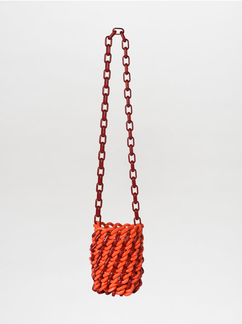 The Anise Bag Red, a small orange woven accessory featuring a striking elongated red chain strap, is showcased elegantly against a plain white backdrop.