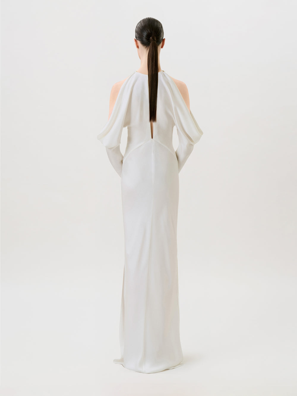 A woman with straight dark hair in a ponytail models the Antonia Dress, crafted in hammered satin and designed with open shoulders. Posed with her back to the camera against a simple light background, she captures the essence of modern ready-to-wear sophistication.