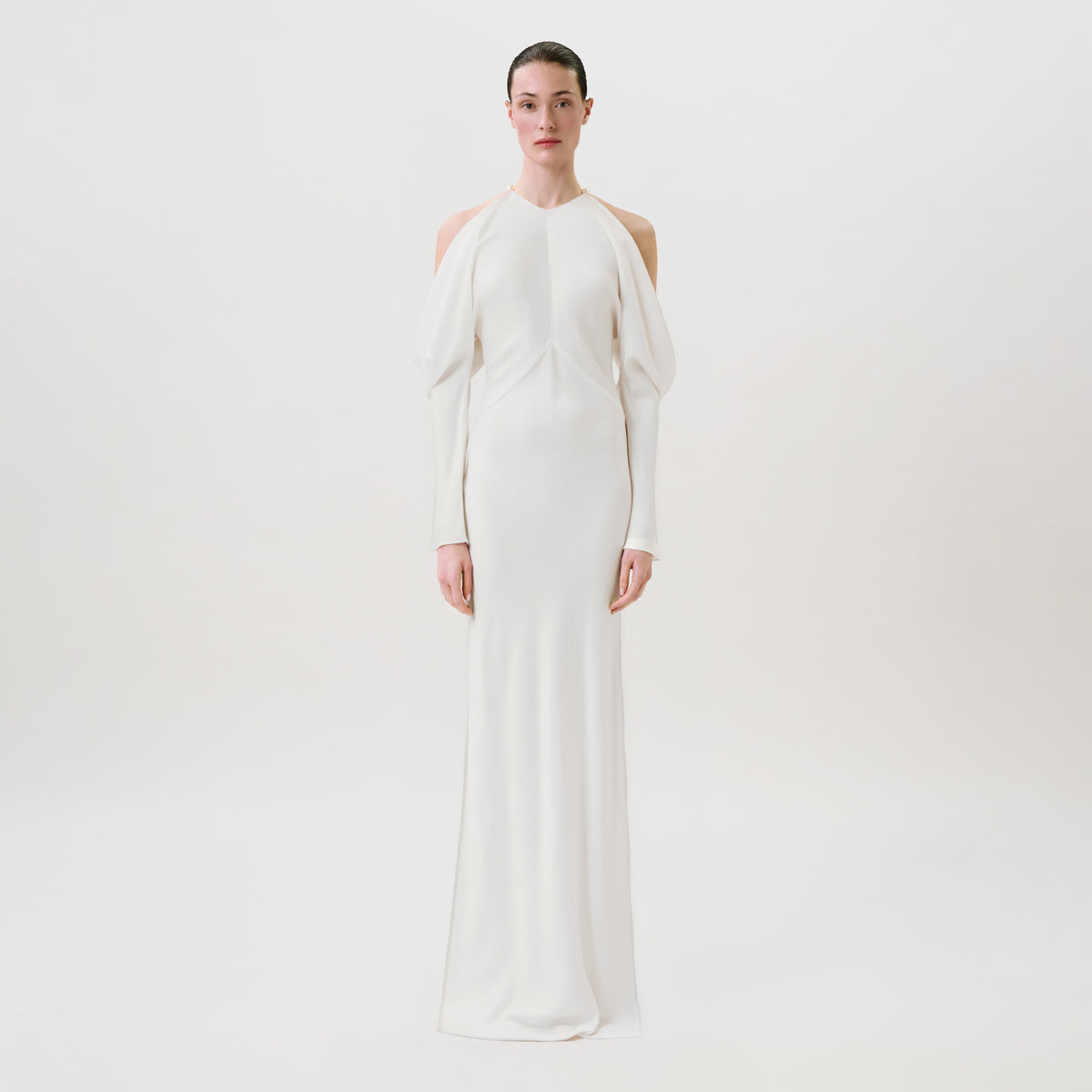 A person stands facing forward wearing the Antonia Dress—an elegant, long white gown featuring cut-out shoulders, long sleeves, and a fitted silhouette crafted from luxurious hammered satin—all set against a plain white background. This Ready To Wear piece exudes modern sophistication.