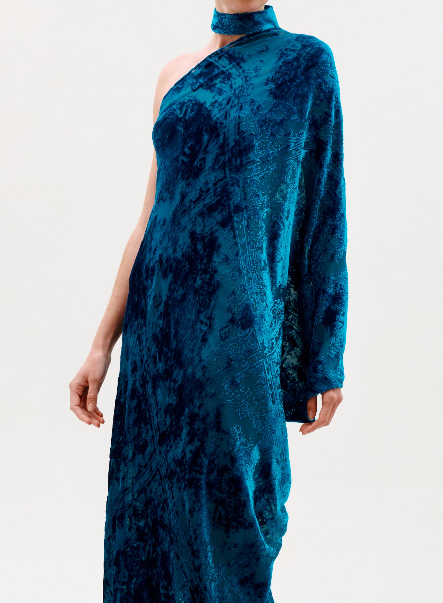 A person models the Anya Dress, a vibrant blue one-shoulder velvet design with a matching choker, single long sleeve, and patterned texture. The plain light background keeps the focus on the dress, with the person's face out of view.