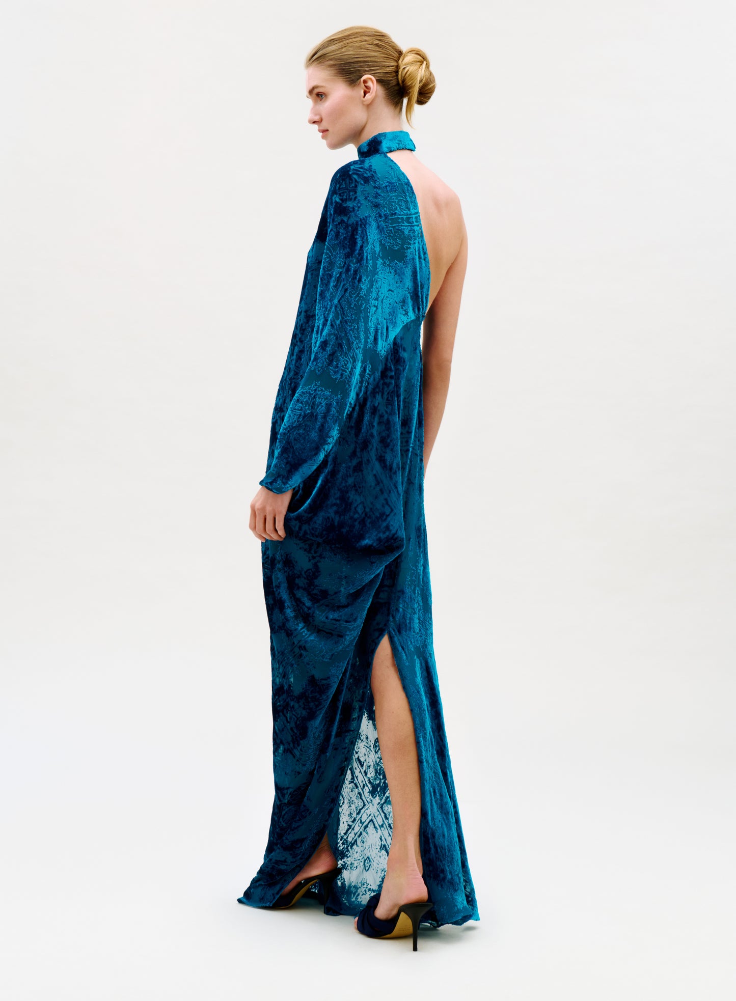 A woman in a low bun showcases the Anya Dress, a long blue velvet gown with an open back and high slit, paired with black heels against a plain light background.