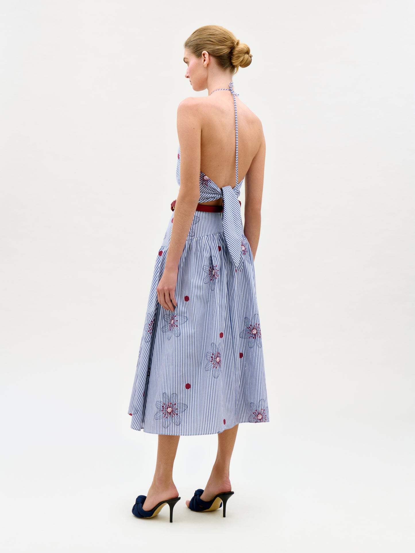 A woman stands on a white background wearing the Ares Dress—a backless, halter-neck, blue-and-white striped midi dress with red floral embroidery, an A-line silhouette, and black heels. Her hair is styled in a low bun.