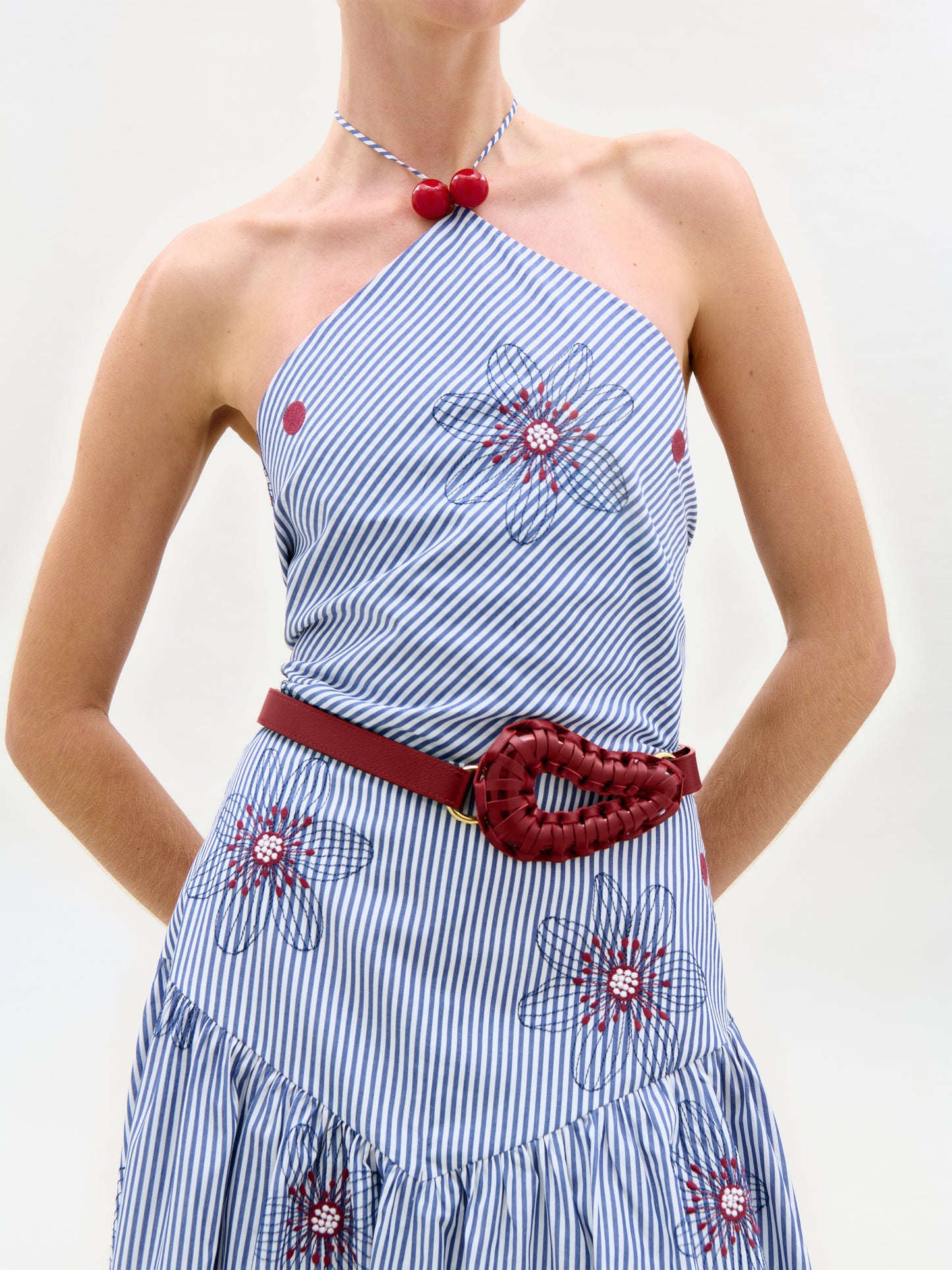 The Ares Dress features blue and white stripes with red floral embroidery, styled with a braided red belt and matching beaded necklace. Its A-line silhouette flatters the figure, shown hands-on-hips against a plain white background.
