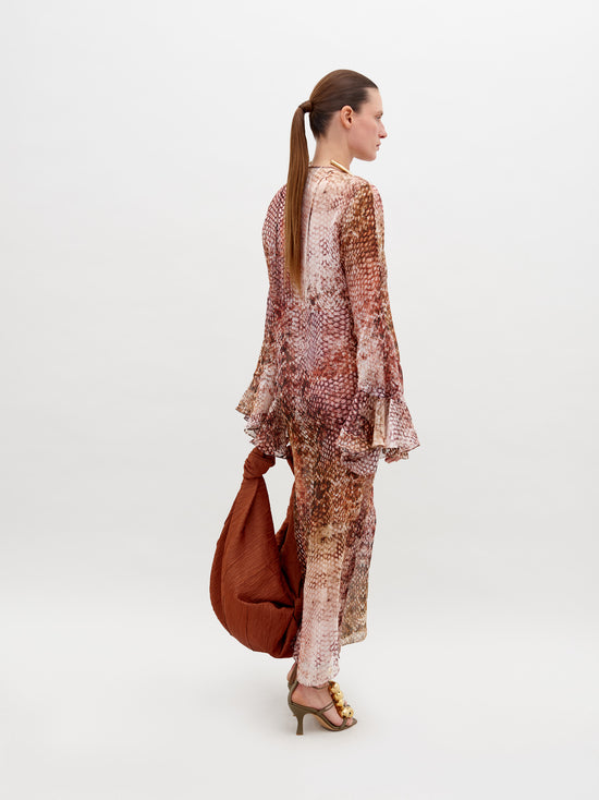 A person stands against a plain background wearing the Aries Dress Peach Animal Print with flared sleeves, embellished accessories, and high-heeled sandals. They hold a large, rust-colored bag. Pre-order now to have it by November 15th, 2024.