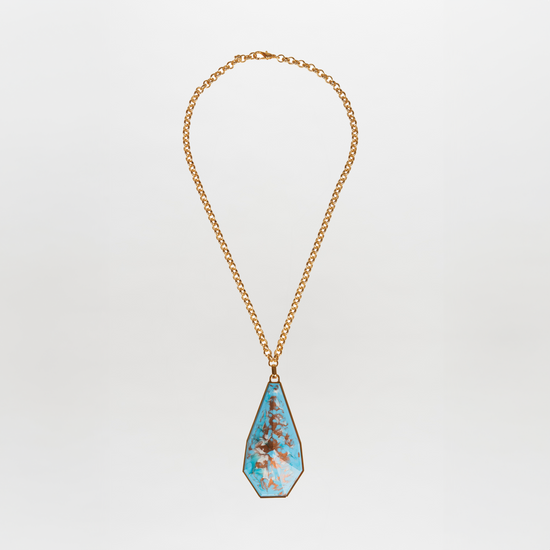 The Ascoli Necklace by Tcherassi showcases a gold chain with a large teardrop-shaped turquoise fantasy stone pendant, accentuated by brown veining and set in an elegant metallic frame against a white background.