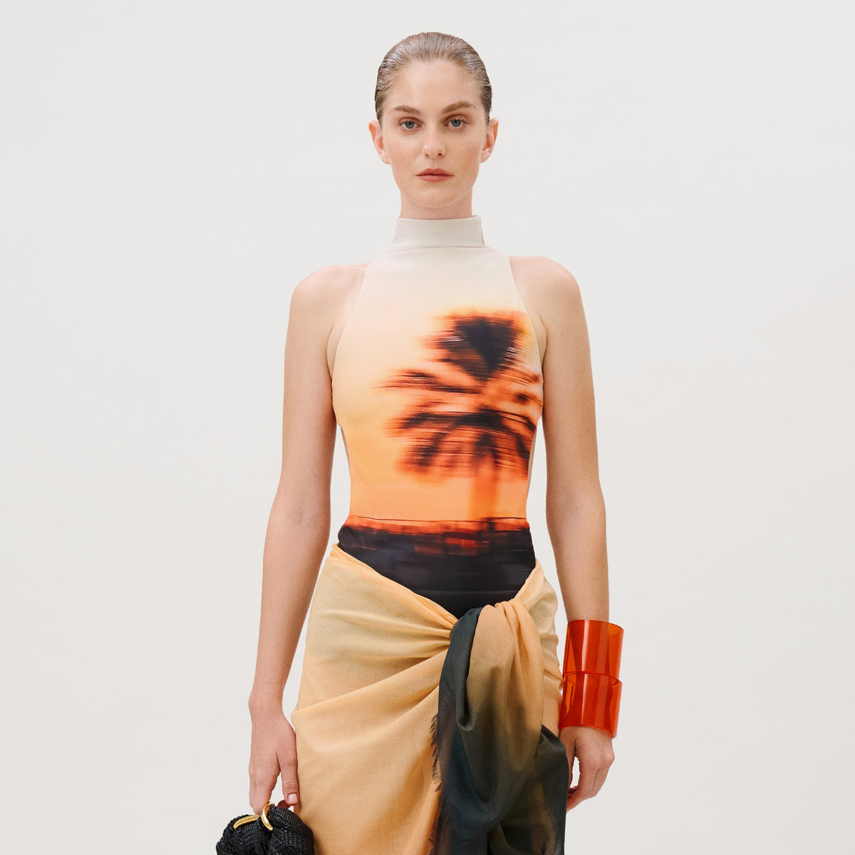 A model wears the Asia Swim Bodysuit, a sleeveless high-neck Lycra piece featuring a sunset and palm tree print, paired with a draped wrap skirt and a large translucent orange bangle.