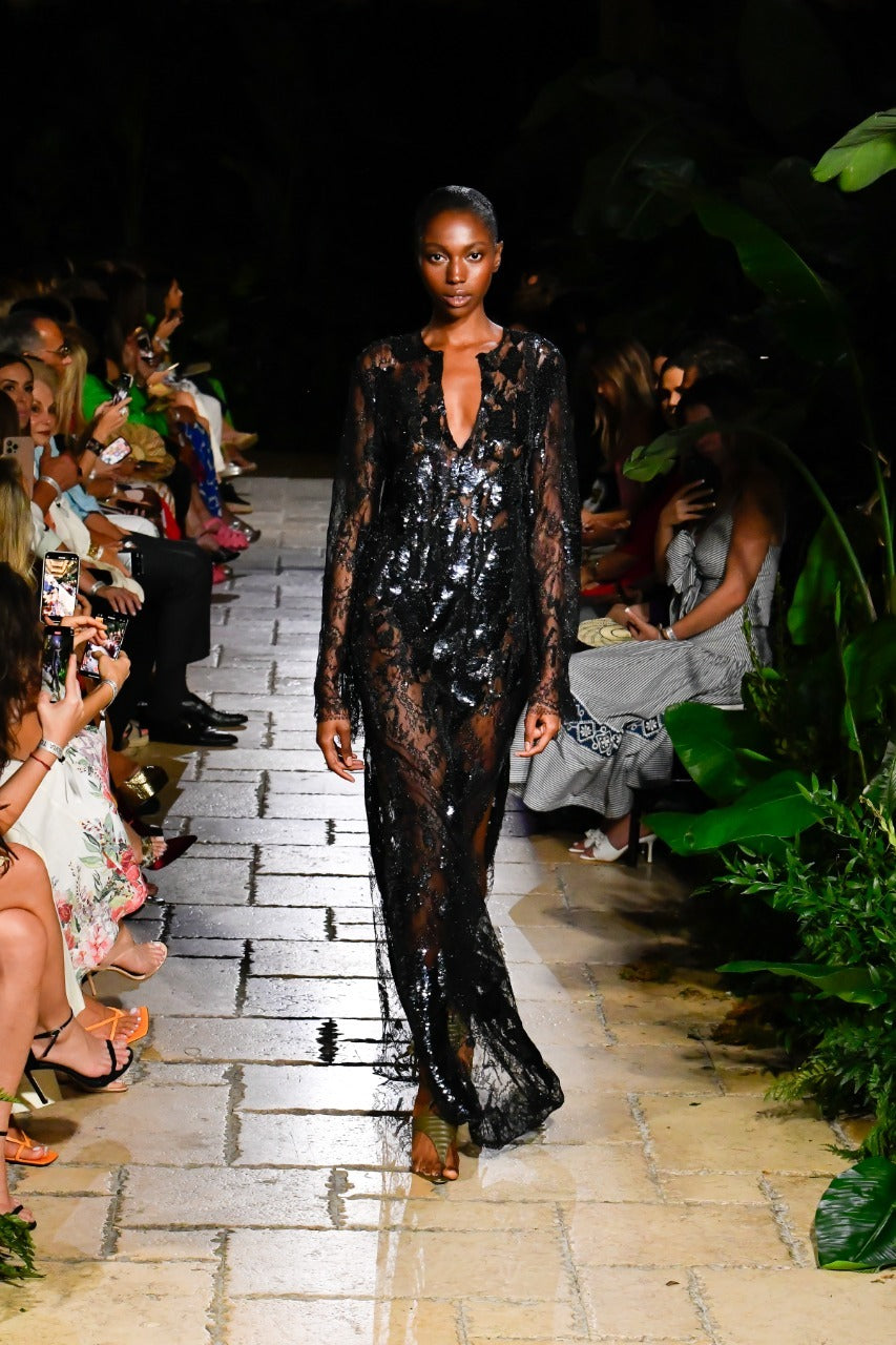 Model walks the runway in a black, sheer, sequined dress. People sit on either side of the walkway, surrounded by greenery.