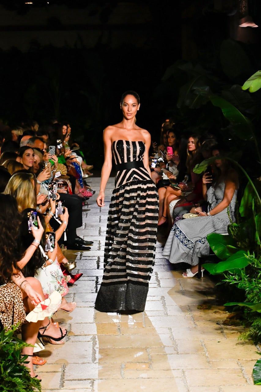 A model walks down a tropical-themed runway wearing a strapless, black and sheer striped gown, surrounded by seated audience members taking photos.