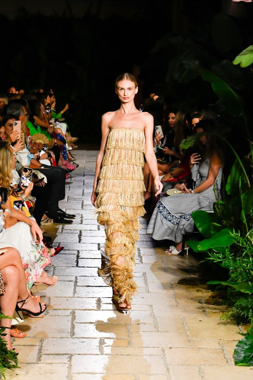 Model walking down a runway in a fringed, strapless dress surrounded by seated audience members in a tropical-themed setting.