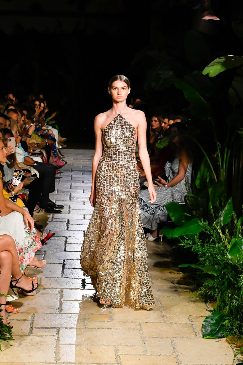 A model walks down a tropical-themed runway wearing a shimmering gold gown, with an audience seated on both sides.