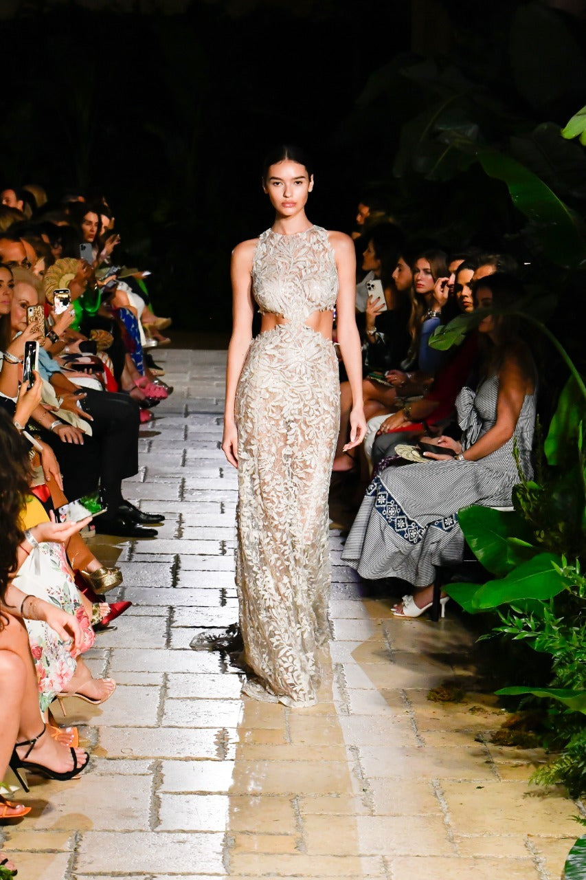 A model walks down a runway wearing an elegant, form-fitting, lace gown. The audience is seated on both sides, observing and taking photos. The setting has lush greenery.