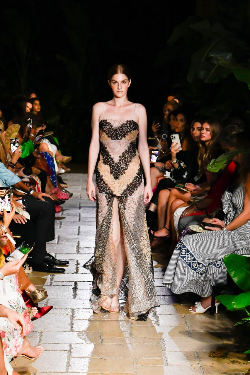 Model walking down a runway in a semi-transparent, patterned dress, surrounded by seated audience on both sides.